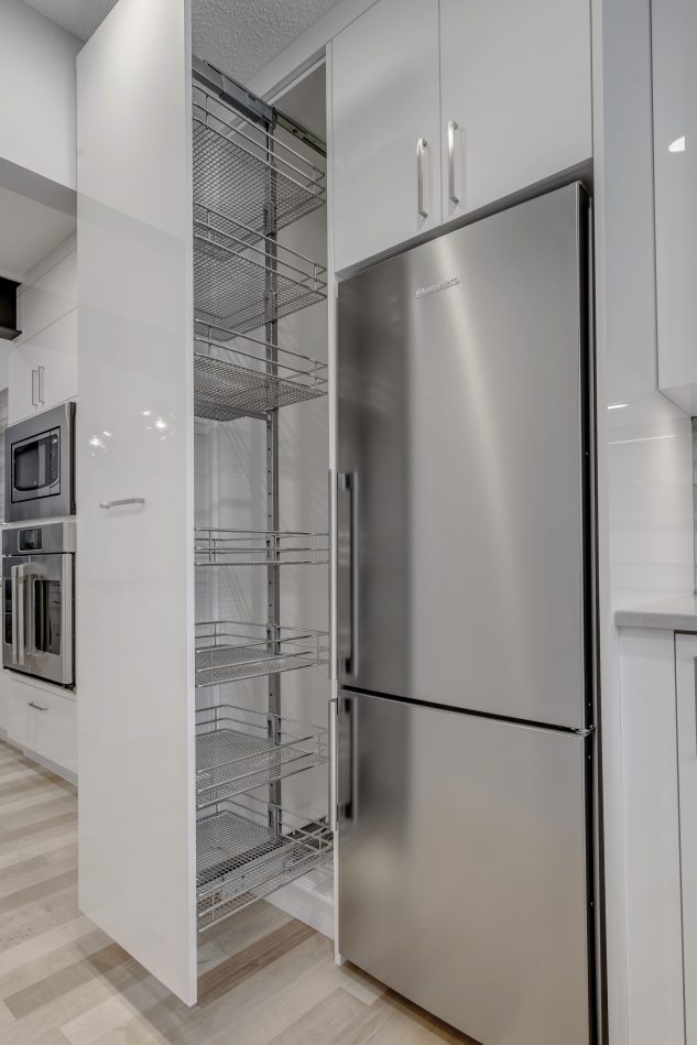 Modern high gloss kitchen cabinetry with grey quartz countertops. Cabinet accessory pull out next to stainless steel fridge.