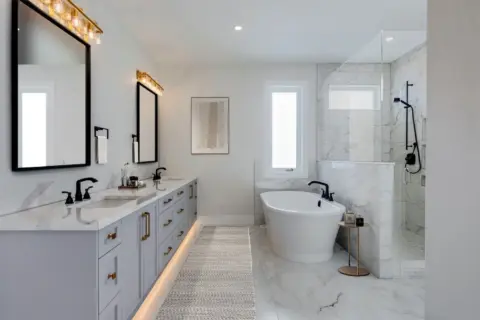 Ensuite with dual sinks, vanity with custom grey Shaker cabinets, quartz countertop, freestanding soaker tub, and expansive tiled shower