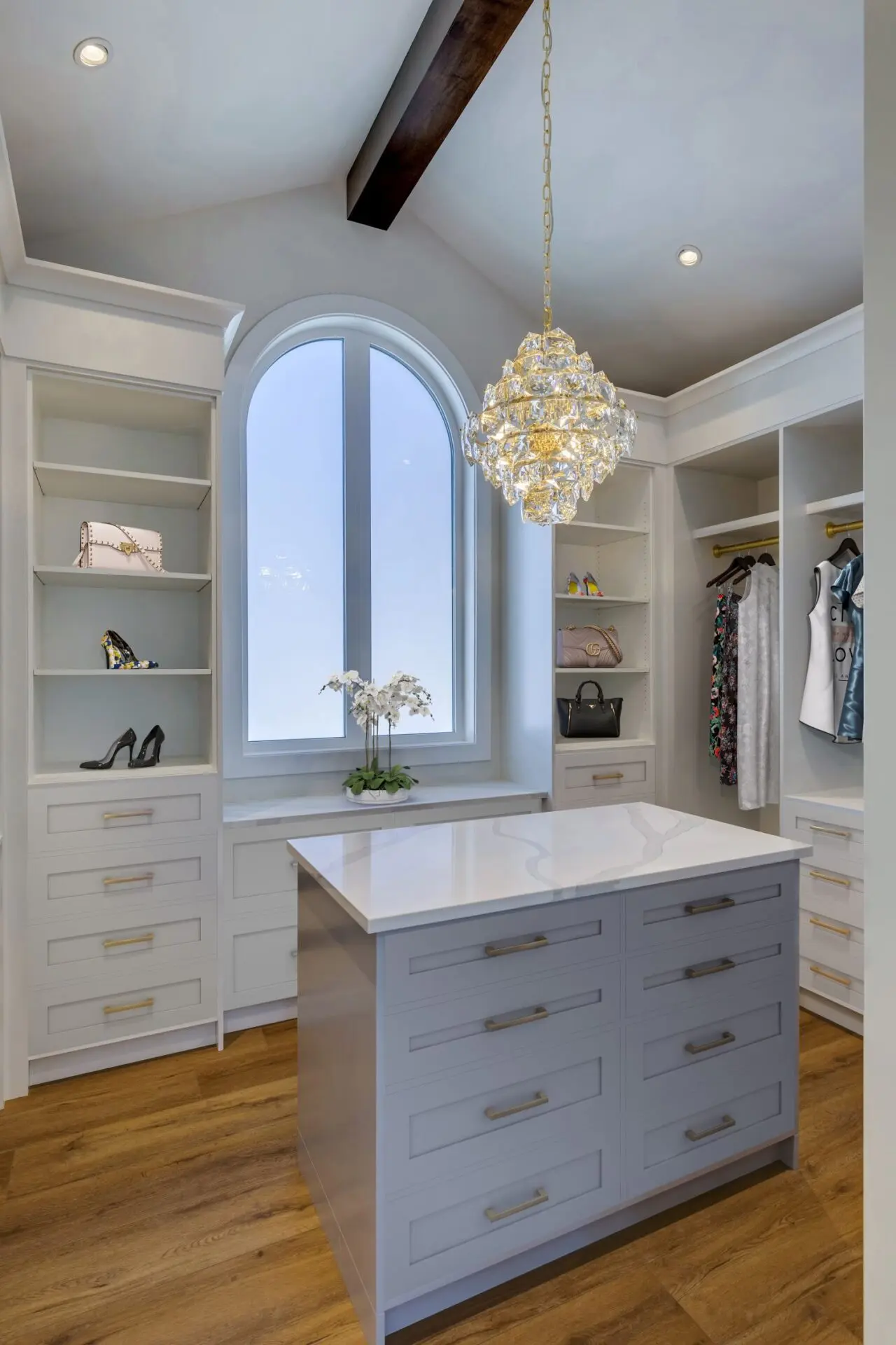 Walk in closet with custom white and grey inset Shaker cabinetry, central island, arched window, chandelier, beam ceiling detail