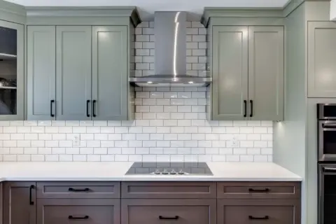 Closeup of two-toned beveled Shaker cabinetry in Louisburg Green and rich custom brown stain with glossy Oyster Gray subway tile in custom Calgary kitchen