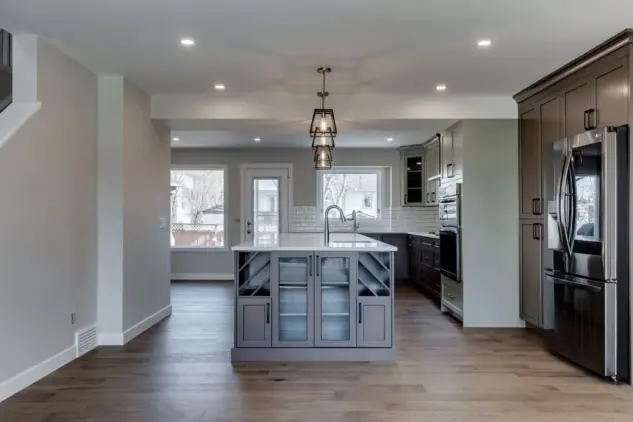 Wide angle view of Calgary kitchen with custom two-tone beveled Shaker cabinets in Louisburg Green and custom rich brown stain, glass front cabinets at end of large island with prep sink