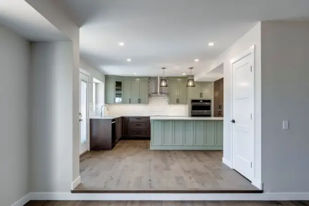 View from family room with one step up to custom Calgary kitchen featuring two-toned beveled Shaker cabinets in Louisburg Green and rich brown custom stain