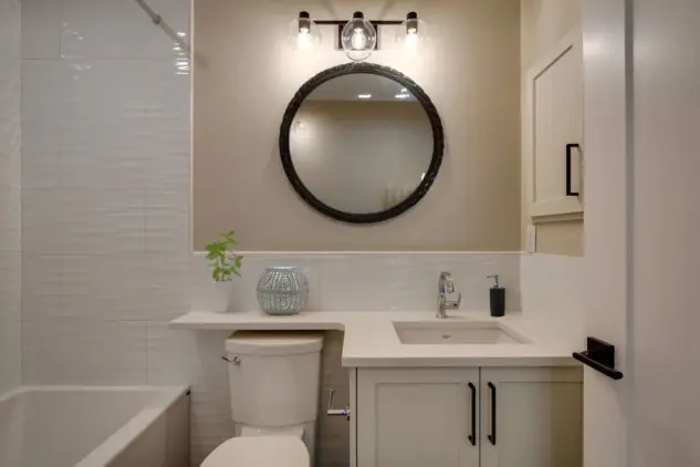 Bathroom in carriage home painted in neutral shade, with tile, quartz countrer and Shaker cabinets in shades of warm white, with chrome tap and black door and cabinetry hardware and black and glass lighting fixture.