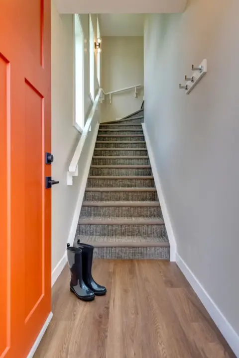 Landing with rich brown LVP flooring and durable carpeted stairway leading up to garage carriage home, and door painted in Benjamin Moore Louisiana Hot Sauce for a welcoming pop of colour.